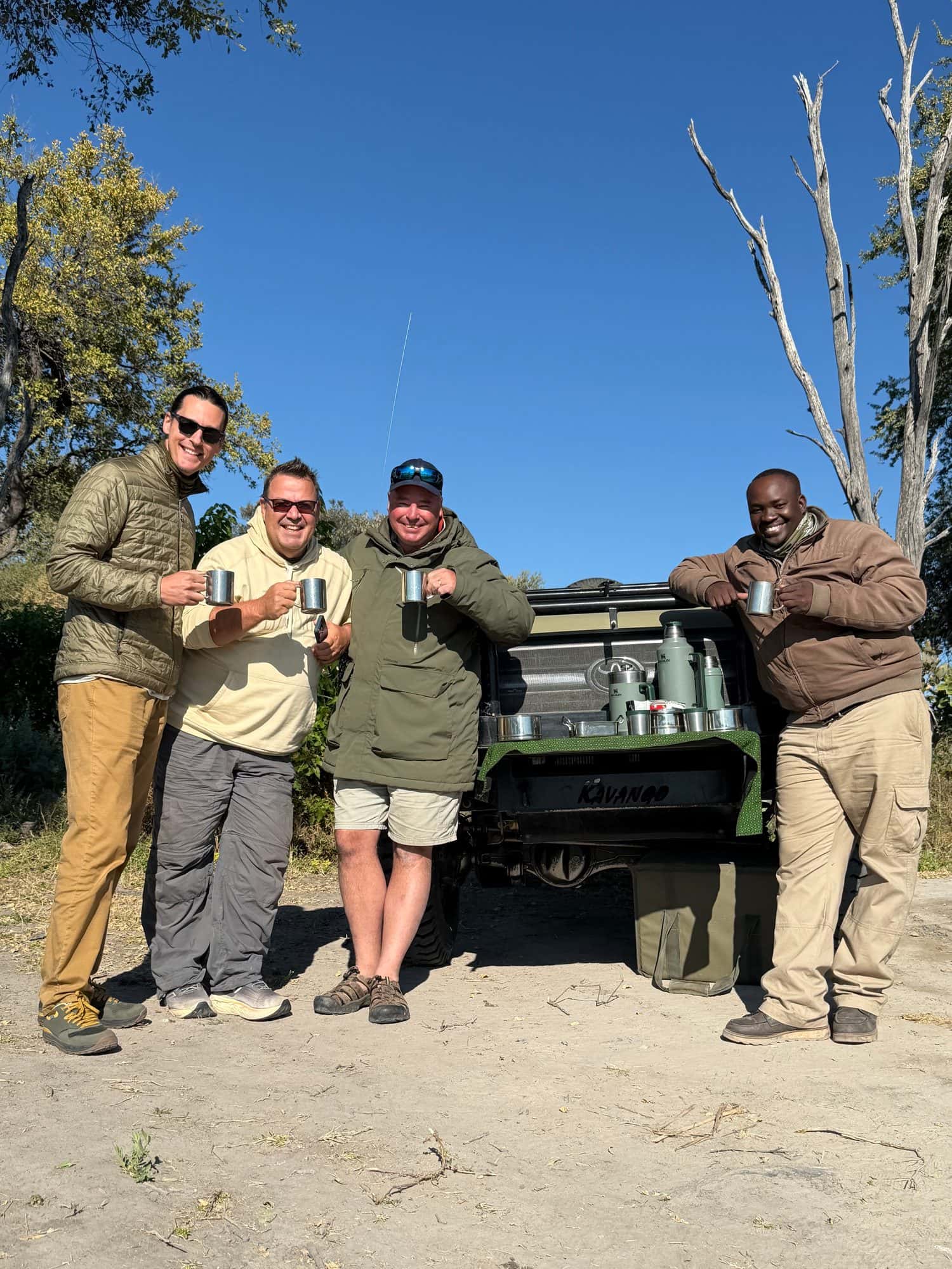 Four men with enamel mugs at the open back of a safari vehicle in the bush