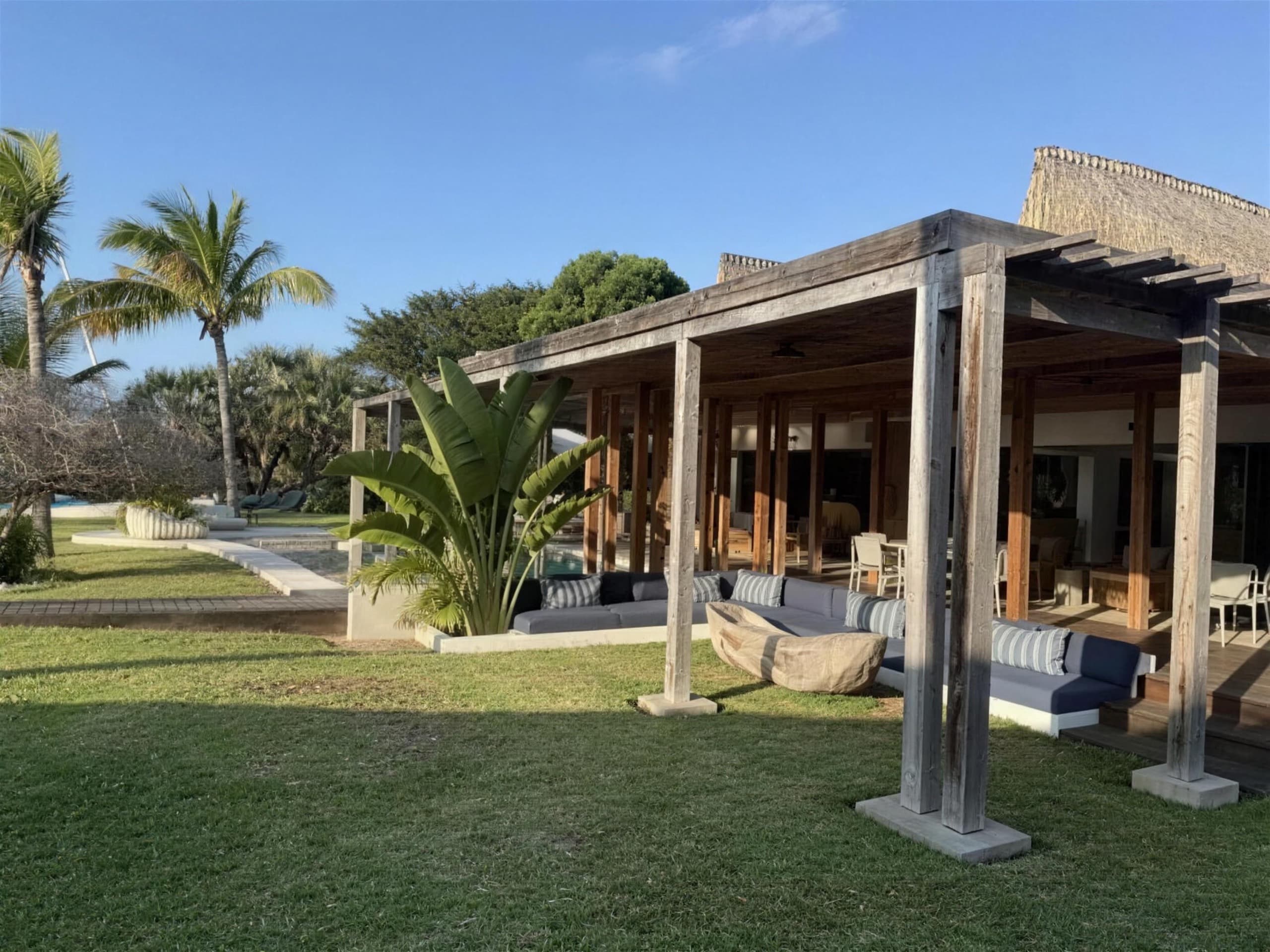 An empty lodge terrace under a timber pergola, with palms and open lawn