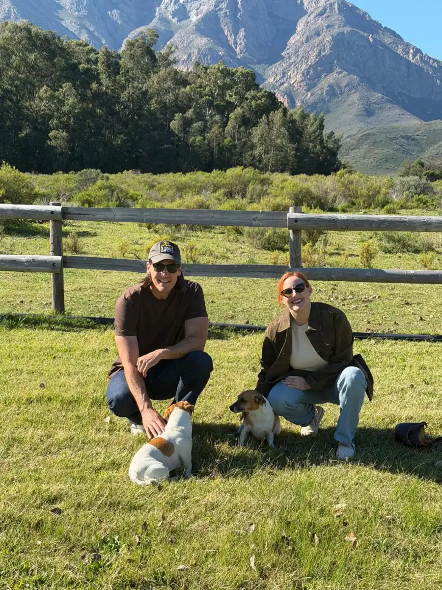 David and Sam Schupp kneeling in the grass with two small dogs, a wooden fence and mountains behind them