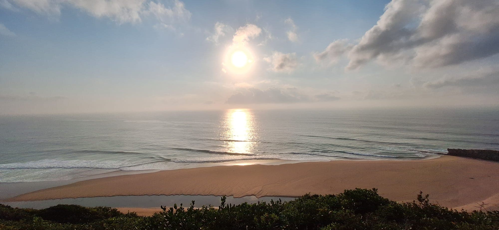 The sun over the sea at Ponta Macumbo, seen from the dune above a wide empty beach where a stream crosses the sand