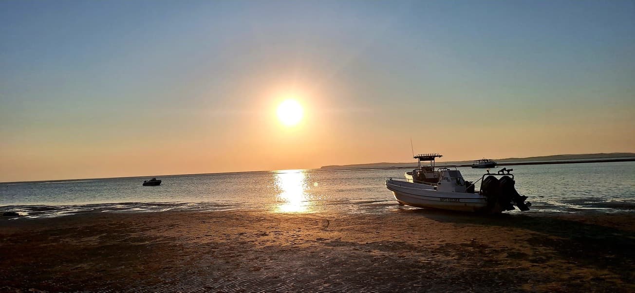 Two boats resting on the wet sand of the bay at low tide, the sun setting over calm water behind them