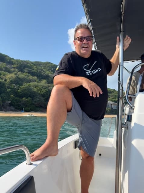 Gordy standing on the bow of a boat with one foot up on the gunwale, green hills and a beach behind him