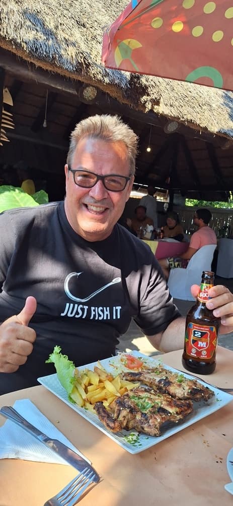 Gordy grinning with a thumbs up behind a plate of peri peri chicken and chips and a bottle of 2M beer under a thatched roof
