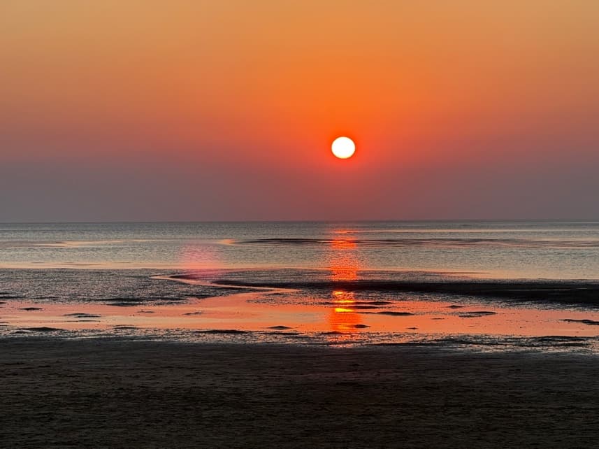 A deep red sun setting over tidal flats, its reflection running across the wet sand towards the camera