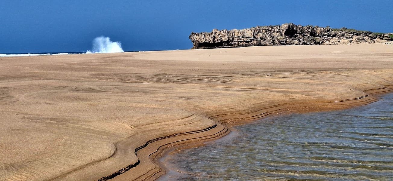 A wide sandbar with a tidal stream cutting through it and a wave bursting against the rocky point behind