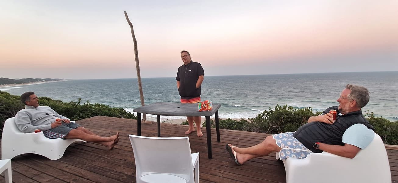 Three guests relaxing on the deck above the sea at dusk, the sky pink over the water behind them