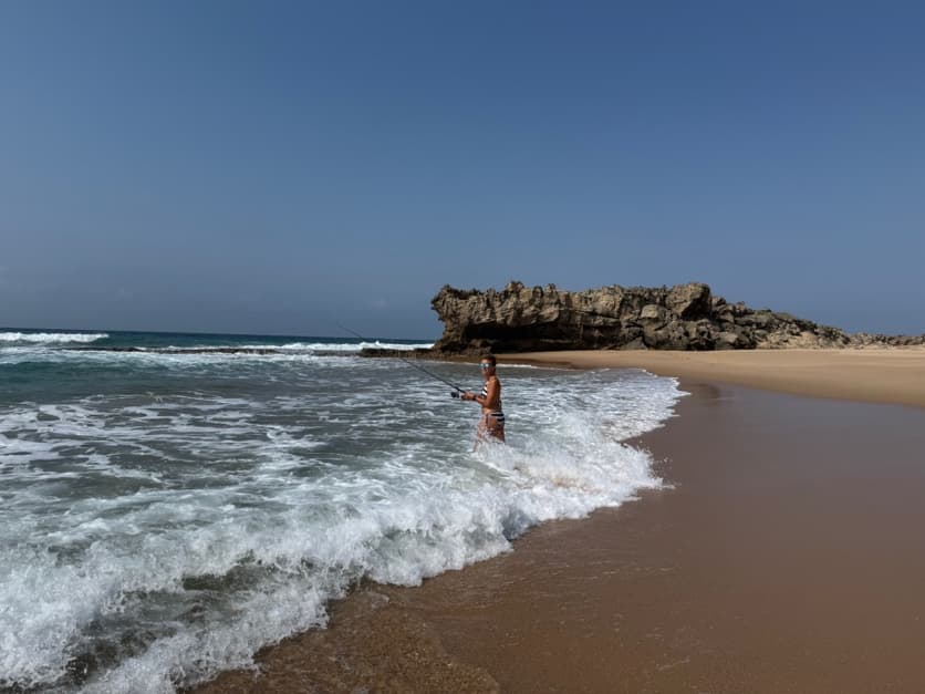 An angler standing in the white water of the shore break, casting towards a rocky point at the end of the beach